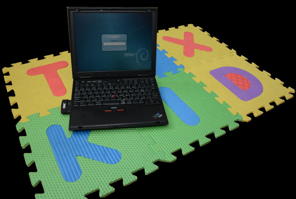Photo of old IBM laptop sitting on mat that reads TUX KID. On the screen is a Debian GUI login prompt